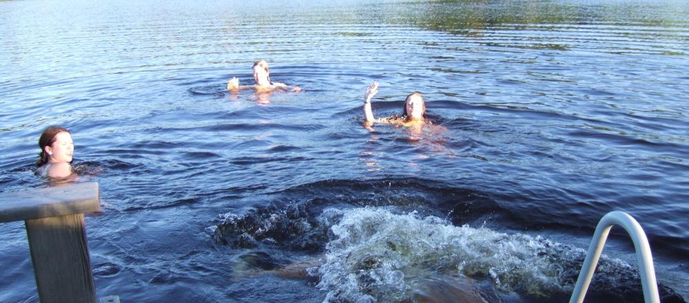 Kolme nuorta järvessä, yksi nostaa kättä tervehdykseksi